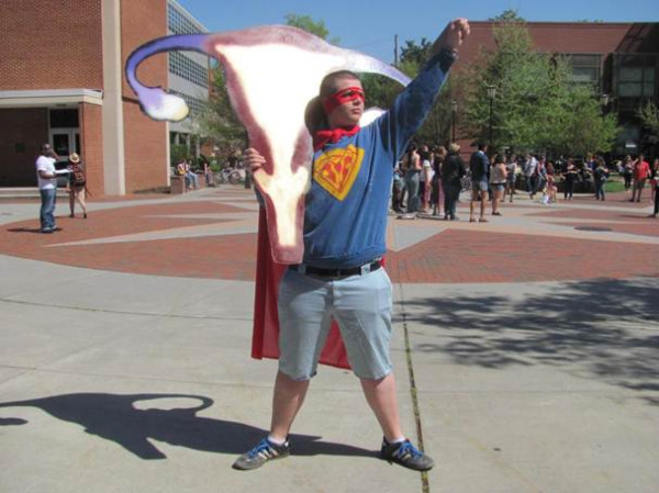A photo of a male student abortion portester dressed like superman except instead of the S on his chest its a picture of the female anatomy and he holding a giant cut out of the female anatomy as well