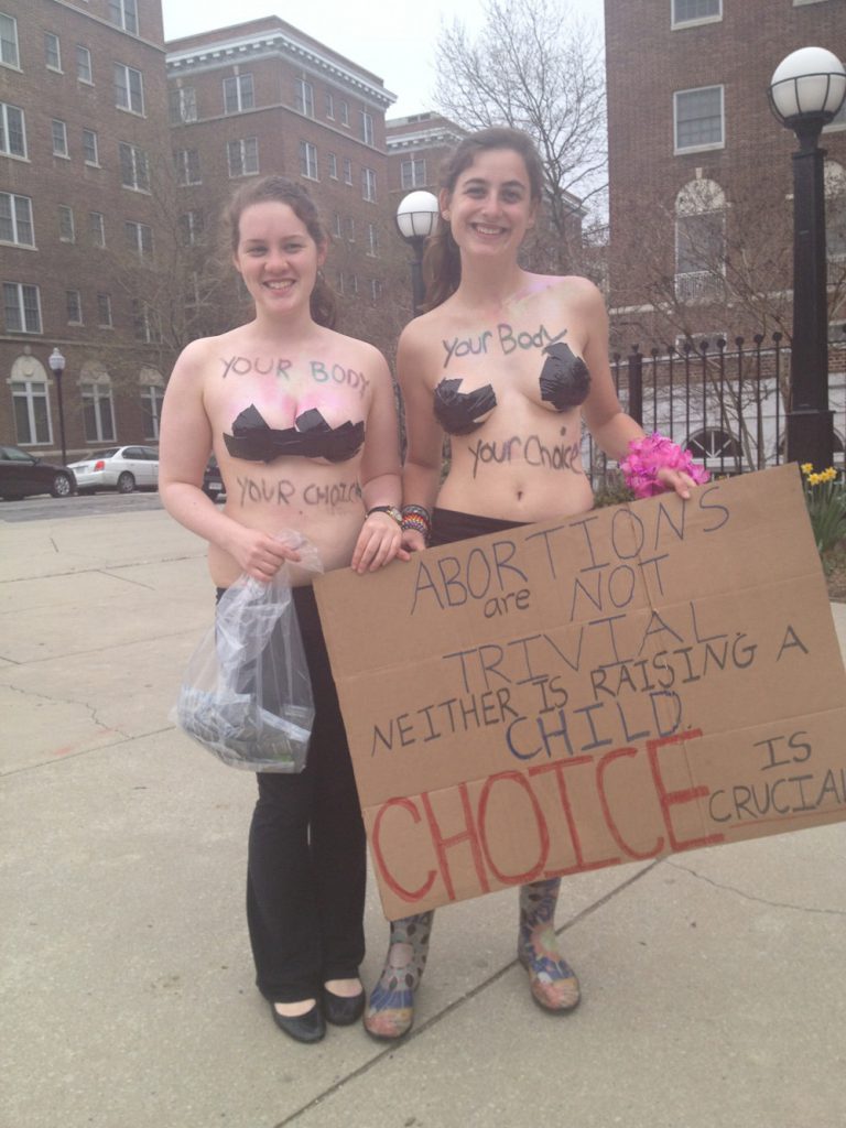 Photo of two topless women (whose breasts are covered with duct tape) with sayings written on their chest and abdomen carrying a sign which reads, "Abortions are not trivial. Neither is raising a child. Choice is crucial."