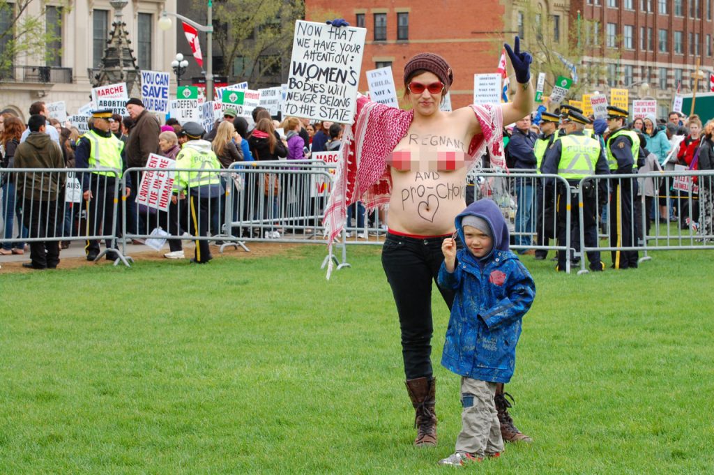 Photo is of a topless pregnant woman with the phrase "Pregnant and Pro-Choice" written on her torso, standing with what appears to be her son and carrying a sign which reads, "Now that we have your attention: Women's bodies will never belong to the state."