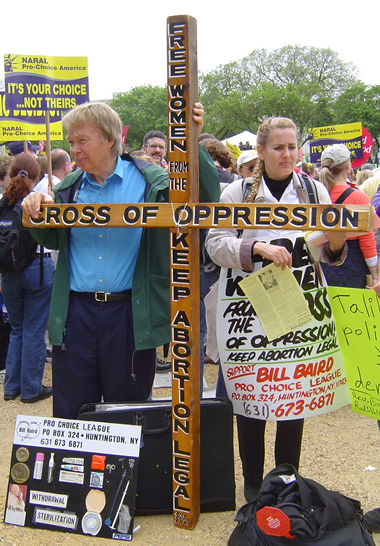 A picture of a male and female abortion supporter holding a wooden cross that reads "FREE WOmen from the Cross Of Oppression keep abortion legal"