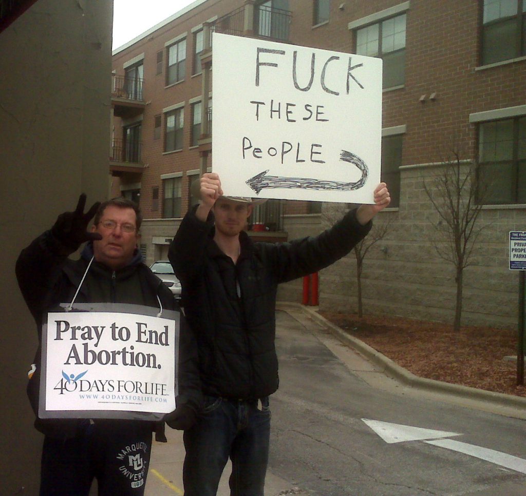 A photo of a male 40 days for life volunteer with a pro-choicer standing next to him with a sign that says: F**K these people