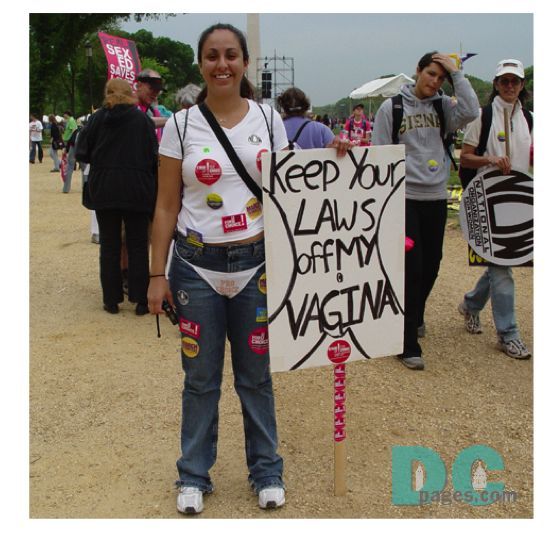 A photo from DC pages.com of a women wearing a white T-Shirt with stickers on it and a pair of underpants over her pants. She also has a sign that says: Keep your laws off my vagina