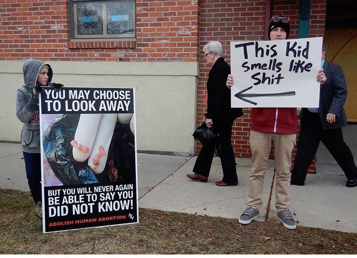 A photo of a young kid holding a graphic abortion sign by Abolish Human Abortion with a slightly older male standing next to him with a sign that says: This kid smells like S**T