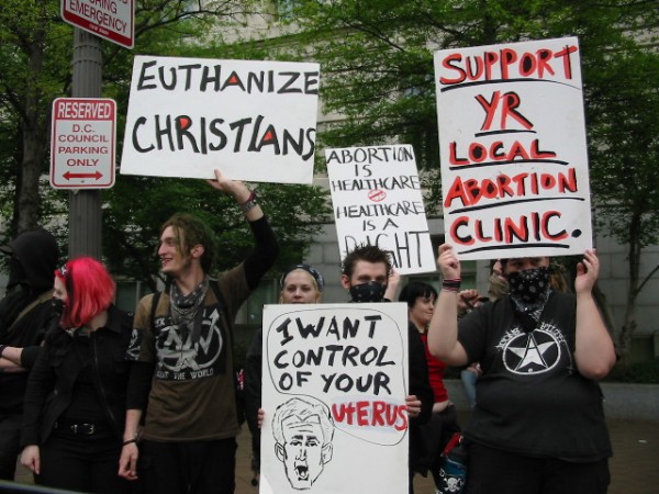 A photo of a group of goth boys holding disturbing signs that say: Euthanize Christians, I want control of your Uterus, Support your local abortion clinic, Abortion is healthcare healthcare is a right