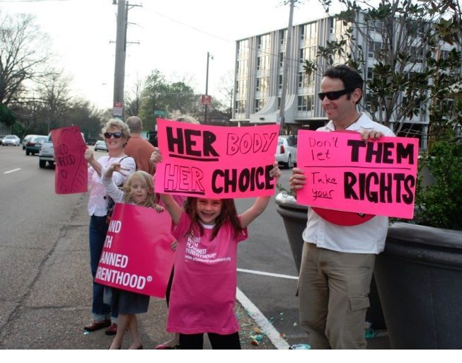 A photo of a Father and his daught both holding signs that read: Her Body Her Choicse and the father is holding the sign that says: Dont let Them take your Rights