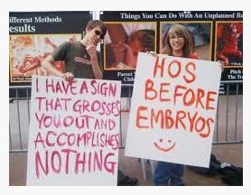 A young male and female are counter protesting a Created Equal graphic abortion display with signs that say: I have a sign that grosses you out and accoumplishes nother, HOS before Embryos :)