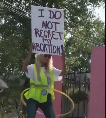 A female clinic escort outside the last abortion clinic in Mississippi hula hooping with a sign abover her head that says: I do not regret my abortion!