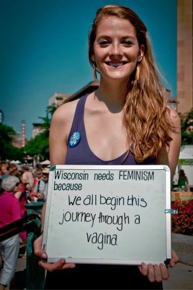 A female in wisconsin holding a sign that reads: Wisconsin needs Feminism because we all begin this journey through a vagina
