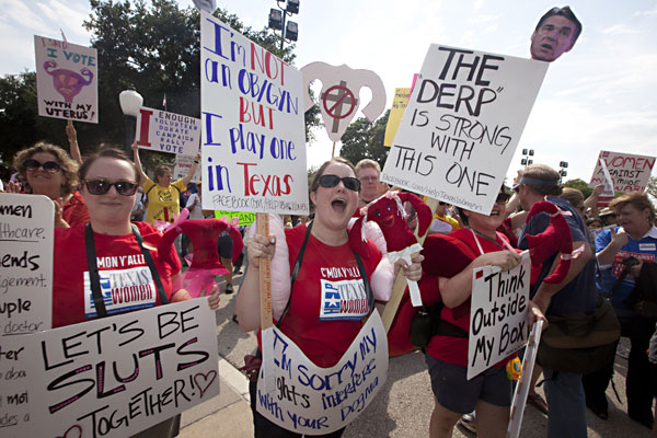 A group of abortion protesters with red shirts that say Help Texas Women on them and holding a stuffed women reproductive system and signs that say: Let's be sluts together!