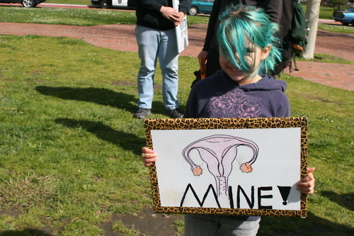 A photo of a little girl holding a sing with a drawing of a female reproductive system on it with the words MINE!