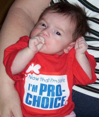 A photo of a mother holding her young baby that is wearing a red t-shirt that says: Now that I'm safe I'm pro-choice