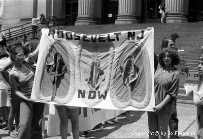 A photo of two women from the Roosevelt NJ chapter of NOW holding a banner with three handmade vagina's on it