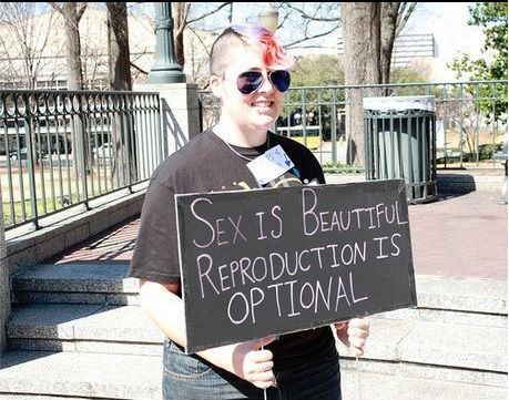 A female with a mohawk holding a sign that says: Sex is Beautiful Reproduction is optional