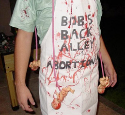 A holloween costume of a bloody abortionist. He's wearing a bloody apron with Bob's Back Alley Abortions on it with naked baby dolls hanging off of it