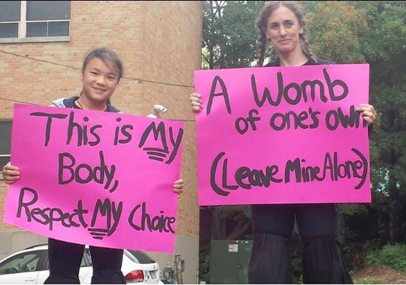 A photo of two girls holding signs that say: This is my body, respect my choice and A womb of one's own leave mind alone