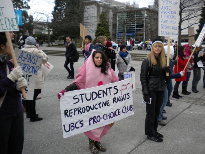 A photo of a young female in a vagina costume holding a sign that says: Students for Reproductive Rights UBC's pro-choice club