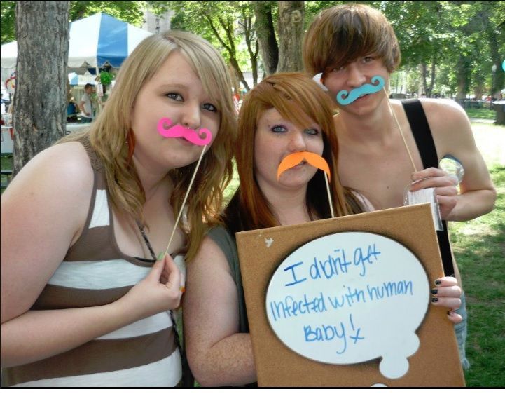 Three youth at a planned parenthood booth. They are holding up colorful mustache's and holding a sign that says: I didnt get infected with human baby!