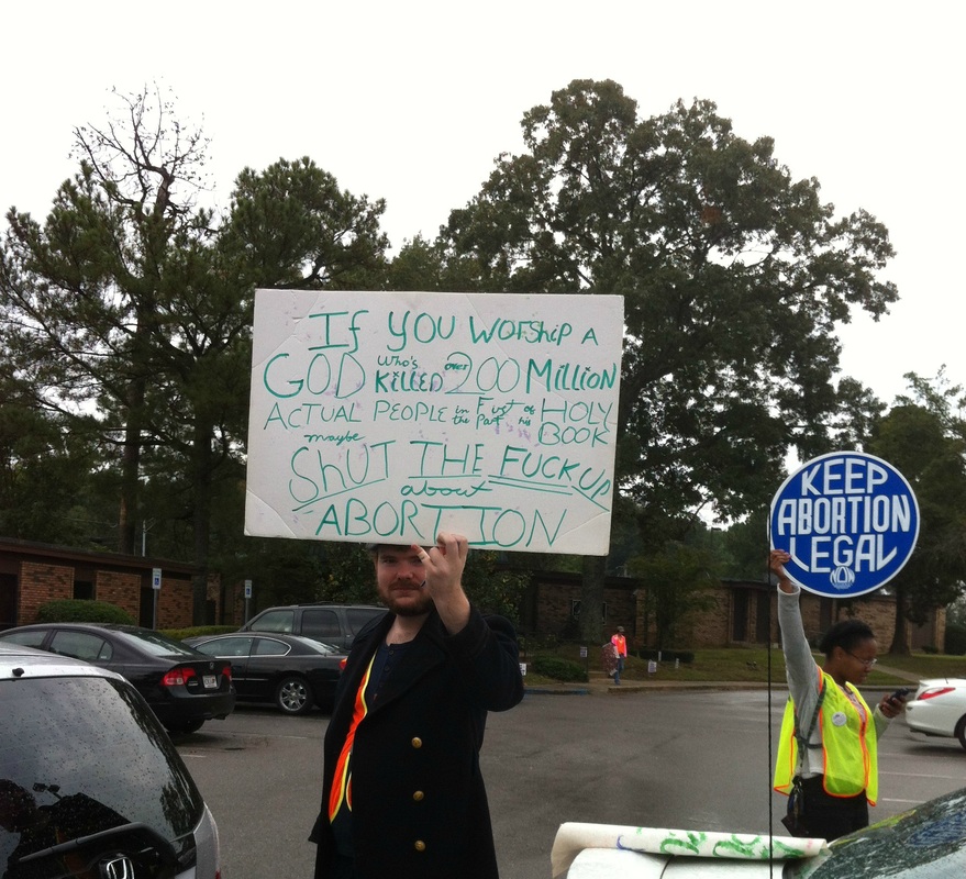 A picture of a male protester standing next to a clinic escort holding a sign that reads: If you worship a GOD who's killed over 200 million actual people in the first part of the holy book shut the F**K up about abortion