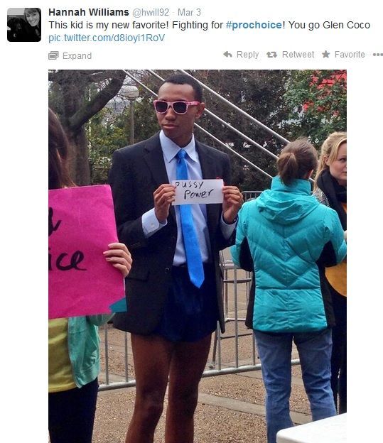 An African American male wearing short shorts a blazer suit shirt and tie holding a small sign that says: P***Y Power