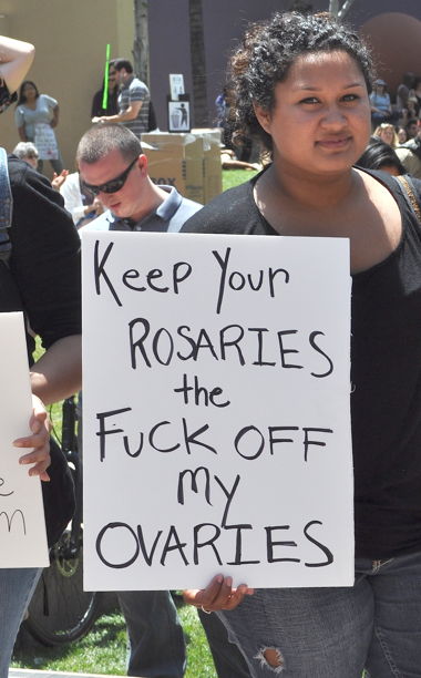 A photo of a female protester holding a sign that says: Keep Your Rosaries The F**K Off My Ovaries