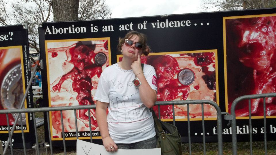 A young female students standing in front of a graphic abortion display, wearing a tshirt that says "This is Rape Culture," pretending to cry about it in a mocking tone.