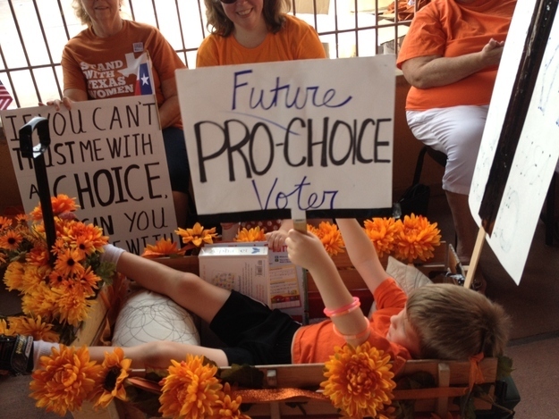 A photo of a young boy laying down in a wagon covered in flowers holding a poster that says: Future PRO-CHOICE Voter