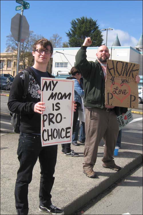 A photo of two males who support abortion with signs that say: My mom is pro-choice and Honk if you love PP