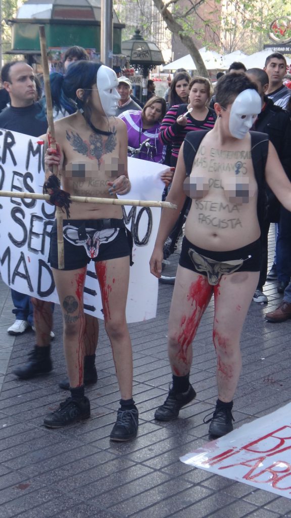 Photgraph taken of two topless females with white maskes on their faces with cut out picture of vaginas with blood streaming down their legs