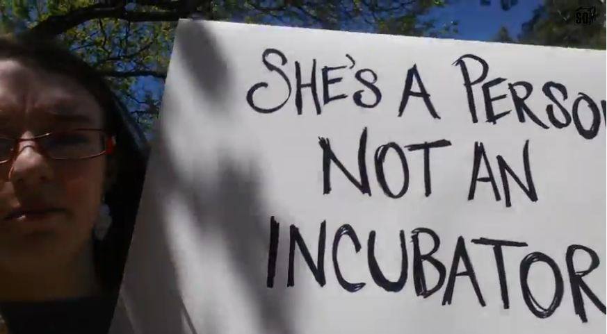 A photo of a femal protester with a sign that says: She's a person not an incubator