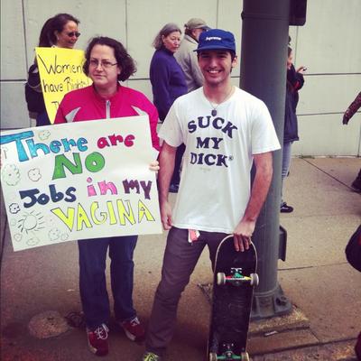 A photo of a Mother and Son at a abortion rally. The mother is holding a sign that says: There are NO jobs in my vagina, and the son has a shirt on that says: suck my d**k