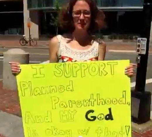 A female protester holding a sign that says: I Support Planned Parenthood, And My God is Okay w/That!