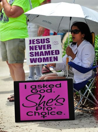 A photo of a women sitting on a sidewalk outside an abortion clinic with two signs that read: "Jesus Never Shamed Women", "I asked God. She's Pro-Choice"