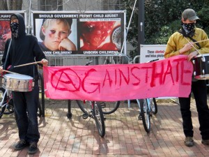 A picture of two students in an anarchist group holding a banner trying to cover up some graphic abortion picture display on campus. Their banner says: Against Hate