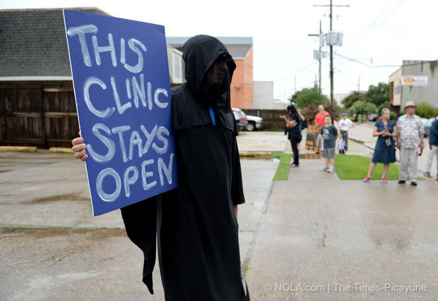 A photo of a protest outside an abortion clinic where a young man is wearing a Grimm Reaper costume holding a sign that says: This Clinic Stays Open