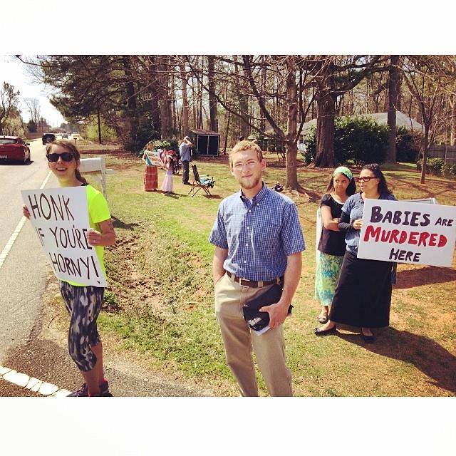 A photo of a group of pro-lifers protesting an abortion clinic, where a husband and wife come and counter protest with random signs that say: Honk If Your Horny!