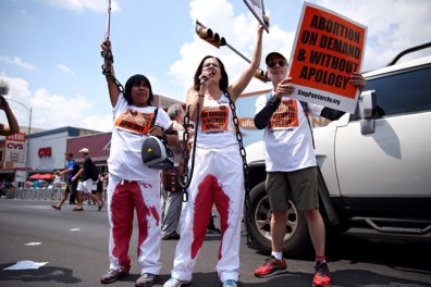 A photo of three members of the abortion rights group Stop Patriarchy are protesting by blocking the street by holding bloody coat hangers and wearing white pants with blood on them as well.