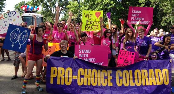 A photo of the group Pro-Choice Colorado at the gay pride parade. Some have signs that say I stand with Planned Parenthood, We Heart everybody's Parts, and even one girl has the femal symbol's next to one another except they are shaped like a vagina
