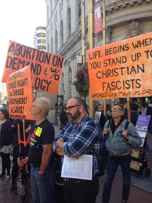 A group of middle age men at an abortion protest next to signs that say: Life Begins When You stand up to Christian Fascists, Abortion on demand and without apology, Demand womens right to choose everywhere! Always!
