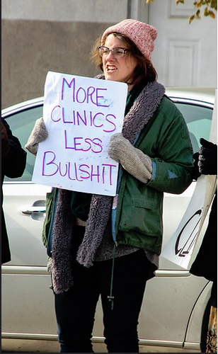 A photo of a young female protesting crisis pregnancy centers with a sign that reads: More Clinics Less Bullshit