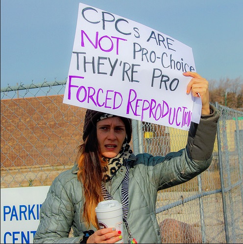 A photo of a female protester holding a sign that says: CPC's are NOT Pro-Choice they're Pro Forced Reproduction