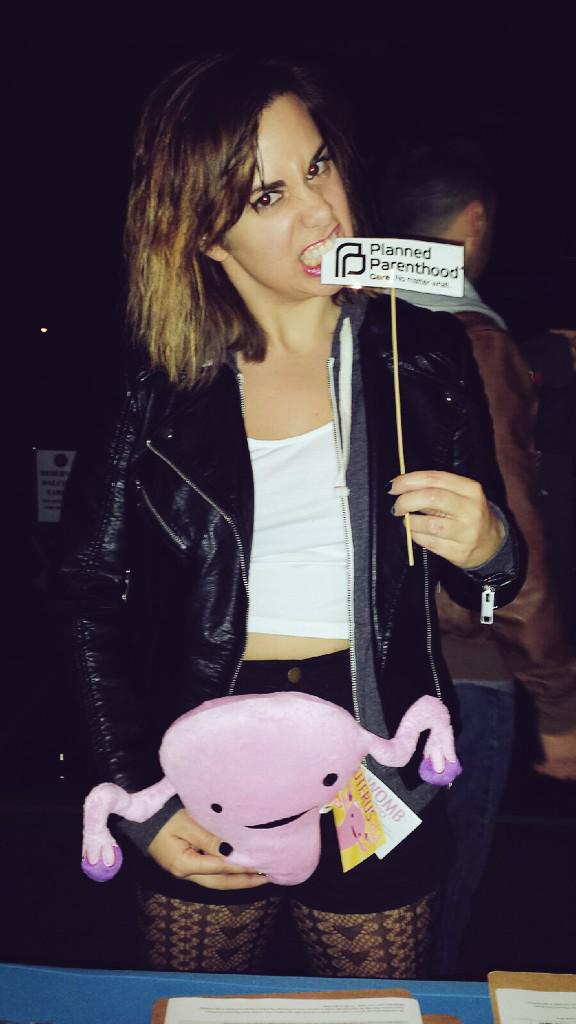 A picture of a female who is biting down on a sign that says Planned Parenthood while holding a pink stuffed uterus toy towards her crotch