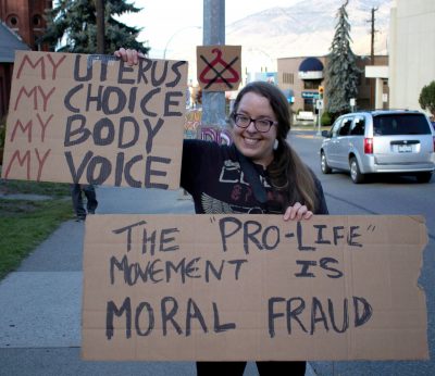 Woman holds two cardboard signs. One reads, "my uterus, my choice, my body, my voice." The other reads, "The 'Pro-Life' movement is moral fraud."