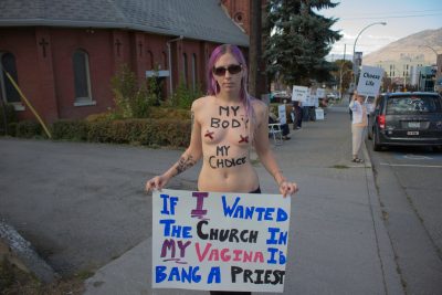 Female protestor wearing no shirt has written on their chest, "my body, my choice" and carries a sign that reads, "If I wanted the church in my vagina I'd bang a priest."