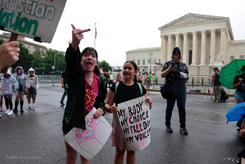 Woman flipping off the camera holds a sign of a woman wearing underwear which reads, "Keep your filthy laws of my silky drawers." Her friend is holding a sign which reads,  "My body, My Choice, My Freedom, My Voice." 