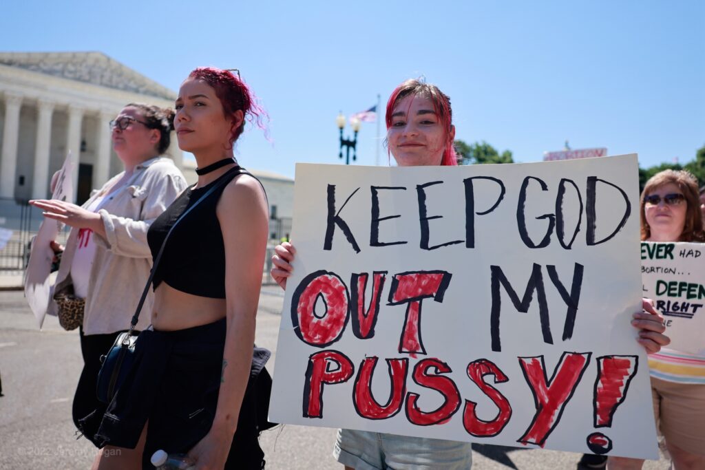 Woman proudly holding a sign which reads, "KEEP GOD OUT OF MY PUSSY!"
